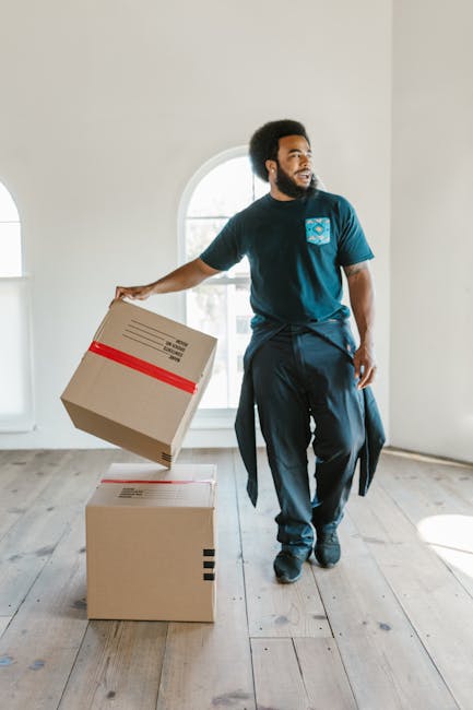A man with curly black hair and a beard is inside a bright, minimally furnished room with wooden flooring and white walls, standing near a large arched window that allows natural light to fill the space. He is wearing a dark blue T-shirt with a logo on the chest, dark pants, and has a jacket tied around his waist. The man is holding a cardboard box with red tape around it in his right hand, lifting it slightly above a second, similarly packed cardboard box placed directly on the floor. The scene depicts the process of home relocation, with the man preparing to load or unload packing boxes, possibly as part of a furniture transport or packing and moving operation. The presence of natural light and the clear view through the window highlight the indoor environment, supported by the professional context of removals or moving services, such as those offered by Man and Van Bloomsbury.