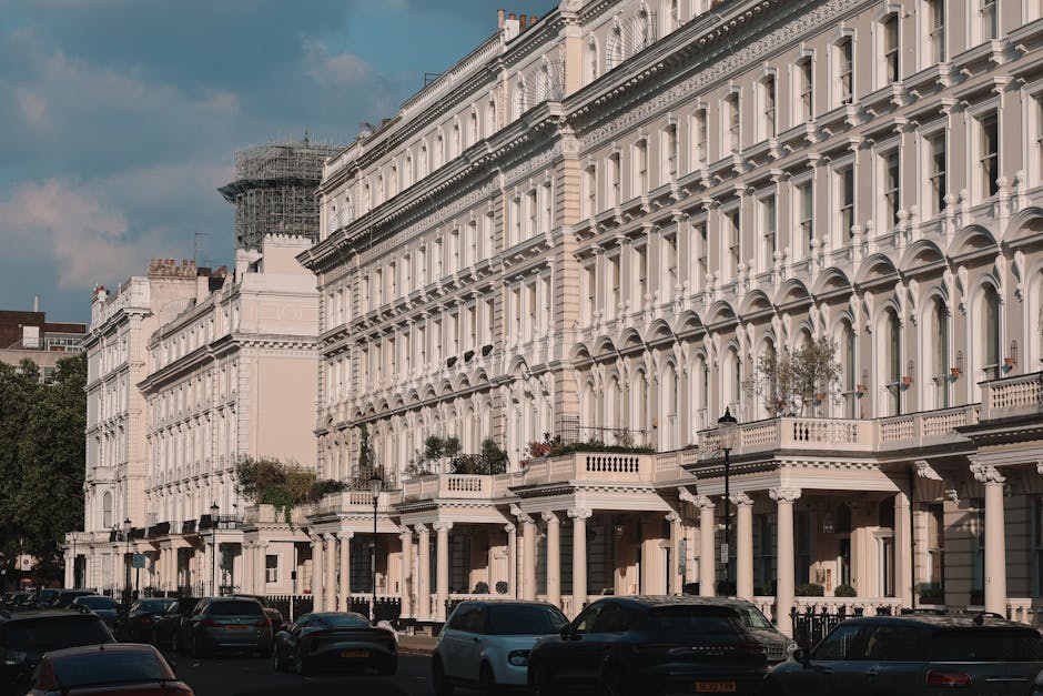 A street scene showing a row of elegant white terraced houses with ornate architectural details, large windows, and decorative balconies, situated behind parked cars along the pavement. The buildings appear to be of Victorian or Georgian style, with some scaffolding visible on one structure, indicating ongoing maintenance or refurbishment. In the foreground, several vehicles, including a black van, a white van, and other cars, are parked parallel to the curb on the asphalt road. The scene is set in daylight with partly cloudy skies overhead, and a lamppost is visible near the front of the houses. This image likely relates to home relocation or furniture transport services offered by Man and Van Bloomsbury, exemplifying the urban environment where professional removals and packing processes are conducted during house moves.