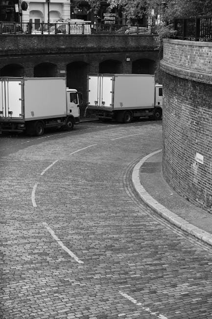 A black-and-white photograph shows three white moving vans parked on a cobbled street near a brick wall and an arched bridge structure, which appears to be part of an urban area with multifloored buildings and trees visible in the background. The vans are positioned parallel to the street and are awaiting loading or unloading. The street features curved white dashed lines indicating parking or loading zones, and the area around the vans is clear of pedestrians. The scene captures a typical home relocation or furniture transport setting during daytime, with the vans likely being used for a moving service such as those provided by Man and Van Bloomsbury, as indicated on the webpage about removals and hidden fees at the specified URL.