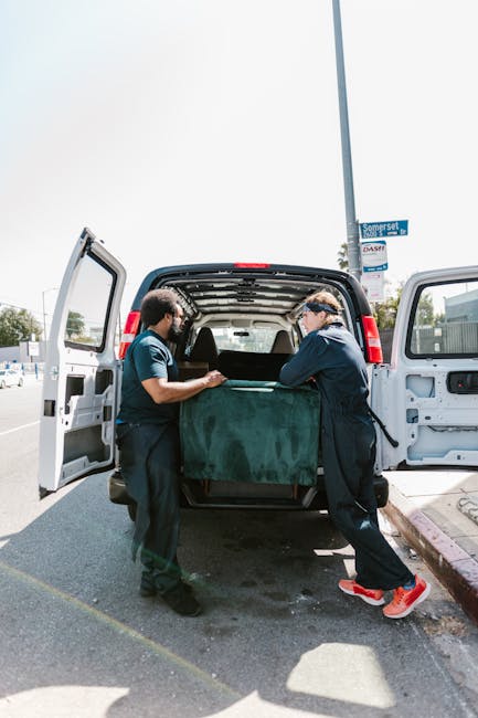 Two professional movers from Man and Van Bloomsbury are loading a large, dark green upholstered piece of furniture into the back of a white cargo van on a paved street. The furniture is partially inside the van, with one mover holding it steady from the left side and the other helping from the right, both wearing dark uniforms and gloves. Inside the van, visible are empty walls and a partially visible plastic strap for securing items during transportation. The scene occurs outdoors in daylight under a partly cloudy sky, with a street sign reading 'Somerset' and a lamppost nearby. The loading process is part of a home relocation or furniture transport service, with the van positioned next to a curb, ready to continue the moving operation. The surrounding area includes parked vehicles and a fenced property, typical of a residential or commercial neighbourhood, illustrating professional packing and moving activities supported by [COMPANY_NAME].