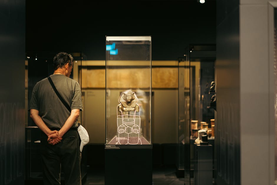 A man, dressed in a gray t-shirt and dark trousers, stands with his hands clasped behind his back inside a museum or gallery, observing a display case containing an ancient artifact resembling a carved stone sculpture of a lion or similar creature. The display case is made of clear glass with a black base, and the artifact is illuminated to highlight its details. The background features dim lighting, dark walls, and other display objects that are partly visible, suggesting a setting focused on historical or archaeological exhibits. This scene reflects a housing or museum environment with the man carefully viewing the preservation of artifacts, and the context aligns with professional furniture transport or packing services for delicate items, such as those offered by Man and Van Bloomsbury as part of their removals and relocation services in the British Museum area.