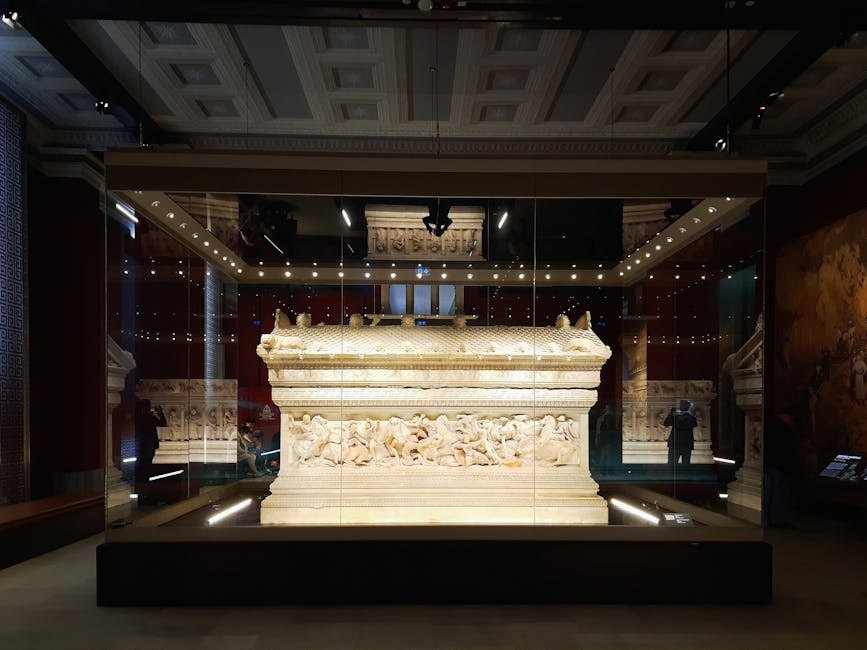 A large, intricately carved stone sarcophagus displayed inside a glass case at the British Museum. The sarcophagus features detailed relief sculptures along its sides, depicting figures and scenes from ancient times. It is positioned on a raised platform within a dedicated display area, illuminated by overhead lighting. Behind the display, the museum interior includes a high, decorated ceiling with recessed lighting, and reflections of visitors can be seen on the glass surfaces. The scene suggests a controlled environment for fragile antiques, with the display prepared for professional handling and transportation moves, such as those provided by Man and Van Bloomsbury for house removals or home relocation involving antique furniture and artifacts.
