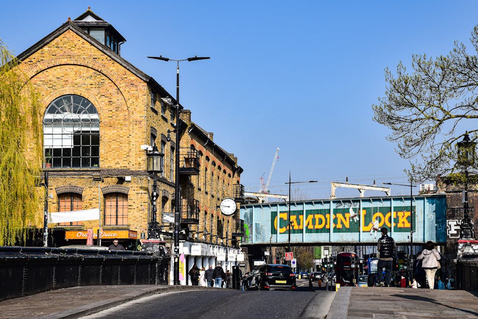 The image depicts a busy street scene in Bloomsbury, featuring a historic brick building with large arched windows on the left, and a prominent green railway bridge with bold yellow lettering reading 'Camden Lock' crossing overhead. Several pedestrians are walking along the pavement, some walking past a small shop with signs for orange juice, while others appear to be waiting or preparing to cross the road. A black van is parked adjacent to the curb, and some people are seen loading or unloading items, possibly related to home relocation or furniture transport. Visible in the background are construction cranes and overhead electric lines, indicating ongoing development. The scene is illuminated by natural daylight, with clear blue skies and a few leafless trees to the right, suggesting early spring. The setting offers insight into street-level logistics and parking considerations in relation to parking regulations for removals, as managed by Camden Council, with Man and Van Bloomsbury services implicitly supporting efficient packing and moving operations in this historic area.
