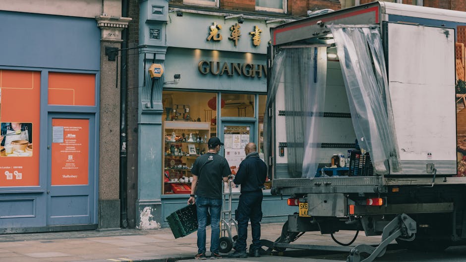 Two men dressed in dark clothing are engaged in a furniture transport and loading process outside a shop with signage displaying Chinese characters and the word 'GUANGH.' They are standing on a pavement adjacent to a large, open-sided moving truck, which is parked close to the storefront. One of the men is carrying a plastic crate, while the other appears to be guiding or discussing the logistics of the unpacking or packing process. The truck, which is equipped with a partially extended side panel and covered with clear plastic wrapping, is positioned in front of the shop, ready for loading or unloading furniture, packaging materials, or boxes. The surroundings include building facades with decorative cornices, and there are some notices or papers posted on the shop window. This scene exemplifies a home or business relocation, highlighting professional moving equipment and coordination typical for a house or office removals service during a packing and moving operation, as conducted by Man and Van Bloomsbury.