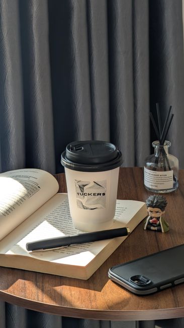 A close-up view of a wooden desk with various objects, including an open book, a disposable coffee cup with a black lid and a logo, a black pen resting on the open pages, a smartphone with a dark case, and a small decorative figure. There is also a glass container with black reed diffusers and a small succulent plant in a white pot. Behind the desk, dark curtains are partially visible, suggesting an indoor setting. The scene appears to be a workspace or home office, with natural light highlighting the objects on the desk, which relate to daily routines or working from home. This setting can be associated with the packing and organizing aspects of home relocations or moves managed by companies like Man and Van Bloomsbury, providing a professional and neutral context for house removals and moving services.