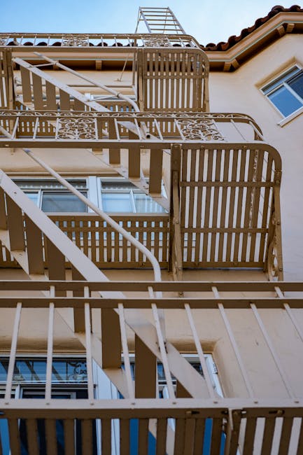 A vertical view of a beige residential building's exterior showing multiple metal fire escape balconies with wooden slats, arranged in a stacked manner on each floor. The fire escapes include curved metal staircases and railings, with some sections covered in protective plastic wrap, indicating recent or ongoing home relocation activities. The top of the building features a terracotta-tiled roof and a metal ladder extending above the fire escape structure. Natural daylight illuminates the scene, highlighting the textures of the building's facade and the metallic and wooden elements of the fire escape. This setting is relevant to furniture transport and packing processes, as companies like Man and Van Bloomsbury efficiently manage removals involving complex stair access and exterior structural considerations.