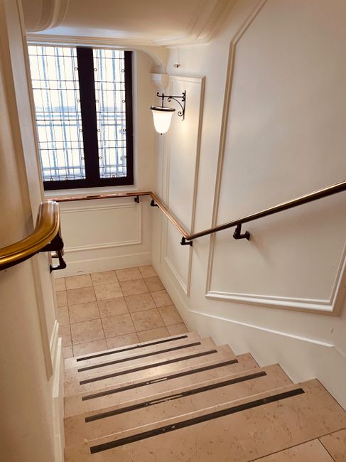 Inside a building stairwell with beige tiled steps and dark anti-slip strips, showing a wooden handrail on the left side and a metal handrail on the right, attached to cream-coloured walls featuring decorative molding. Near the top of the stairs, a black-framed window with diamond-patterned leaded glass allows natural light to illuminate the space. A wall-mounted sconce with a frosted glass shade provides additional lighting. The stairwell appears clean and well-maintained, with neutral tones creating a professional environment suitable for house removals and furniture transport. The image represents an interior space often involved in the logistics of home relocation, where careful handling of furniture is necessary to navigate the tight stair access, as managed by a specialist like Man and Van Bloomsbury.