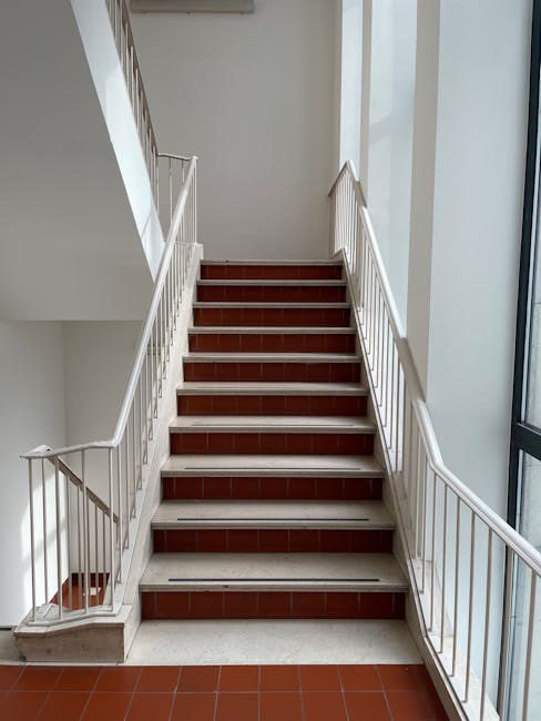 A vertical view of a beige residential building's exterior showing multiple metal fire escape balconies with wooden slats, arranged in a stacked manner on each floor. The fire escapes include curved metal staircases and railings, with some sections covered in protective plastic wrap, indicating recent or ongoing home relocation activities. The top of the building features a terracotta-tiled roof and a metal ladder extending above the fire escape structure. Natural daylight illuminates the scene, highlighting the textures of the building's facade and the metallic and wooden elements of the fire escape. This setting is relevant to furniture transport and packing processes, as companies like Man and Van Bloomsbury efficiently manage removals involving complex stair access and exterior structural considerations.