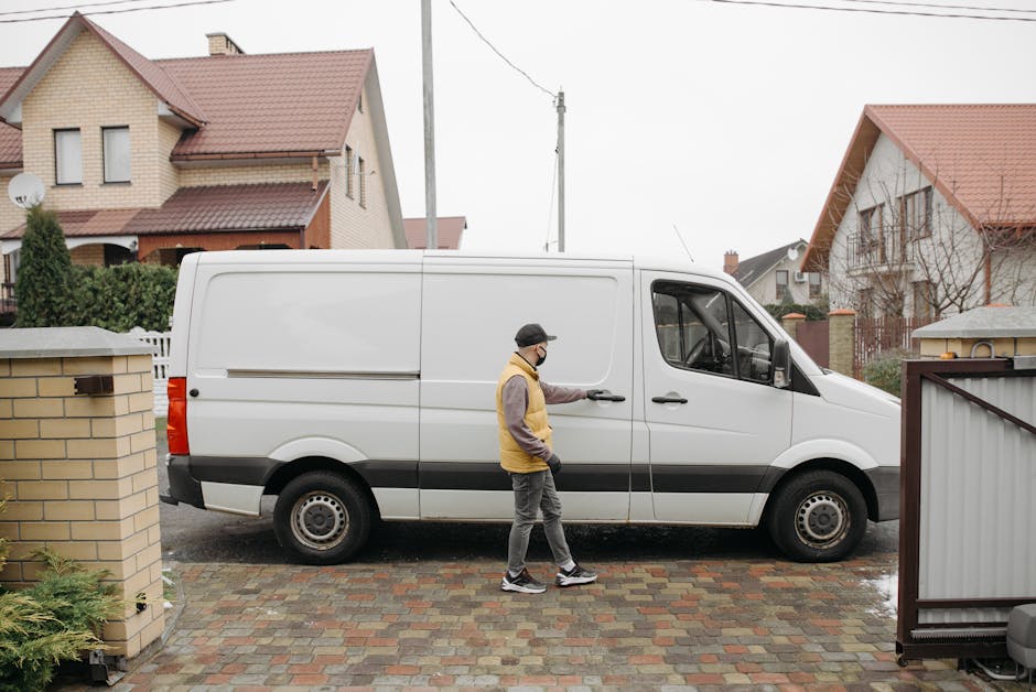 A worker from Man and Van Bloomsbury is seen loading a large number of cardboard boxes into the open rear doors of a moving van parked outdoors on a city street. The boxes vary in size and are stacked