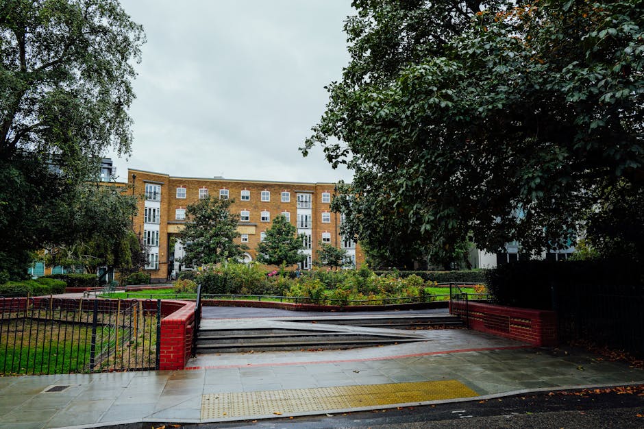 Exterior view of a residential building situated behind a public park with a pathway and garden beds. The pathway leads to a set of short stairs with black metal railings, which are partially blocked by wooden boards, indicating a temporary access restriction. The building has a multi-storey brick facade with white-framed windows and balconies. Surrounding the scene are large, leafy trees providing partial shade. In the foreground, a paved pavement runs parallel to the park, featuring tactile paving for accessibility. The overcast sky suggests diffused lighting. This setting reflects a typical urban environment where house removals and furniture transport might encounter access challenges, which companies like Man and Van Bloomsbury aim to address with appropriate solutions during home relocation or moving processes.