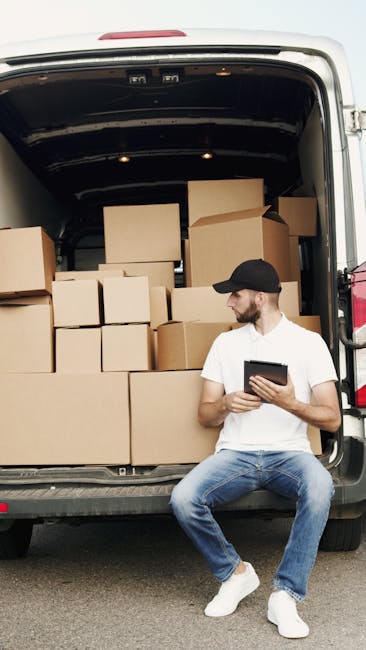 A man wearing a black cap, white t-shirt, and jeans is sitting on the pavement next to the open rear doors of a van loaded with numerous cardboard boxes of various sizes, some sealed with packing tape and others open partially revealing packaging materials inside. The boxes are stacked inside the van in an organized manner, filling much of the cargo space which has a dark interior roof. The man is holding a tablet, possibly coordinating or reviewing the moving process, while the van is parked outside a property, ready for home relocation or furniture transport as part of a professional removals service. The scene reflects the packing and loading stages of a house move, with the boxes prepared for transportation by [COMPANY_NAME] during furniture transport and moving logistics. The environment is outdoor with natural lighting, capturing a moment in the loading process of a house removal.