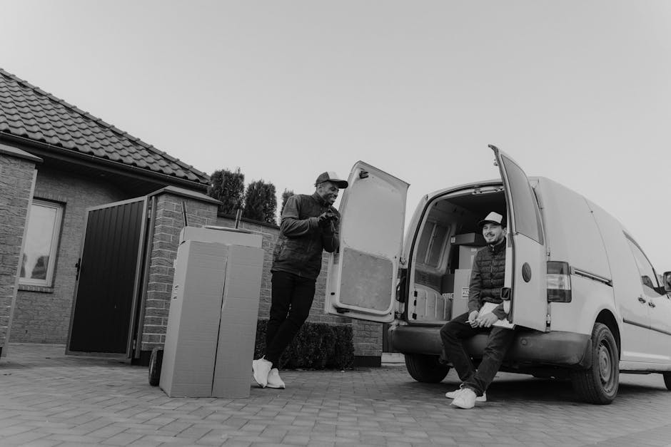 Two men engaged in home relocation activities outside a residential property with a tiled roof and brick exterior. One man is standing on the paved driveway, holding a smartphone, dressed in casual clothing and white sneakers. The second man is sitting on the open back door of a white van used for furniture transport, wearing a cap, a puffer jacket, and jeans, smiling at the camera. The van’s internal space shows various cardboard boxes and packing materials, including plastic-wrapped items and padding blankets, indicating packing and moving preparations. A large cardboard box and a black storage unit are positioned nearby on the pavement, ready for loading. The scene captures the loading process, with the van positioned close to the house for efficient furniture and box transport, illustrating elements typical of house removals and packing activities. Man and Van Bloomsbury’s services are relevant to this context, focusing on home removals and transport logistics.